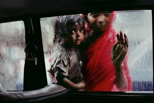 INDIA-10214, Bombay, India Mother and Child at Car Window, Bombay/Mumbai, India, 1993 A mother and child beg for alms through a taxi window during the monsoon. Bombay is the capital of India's business, movie, music, and fashion worlds. A city of wealth, but everywhere, within a few steps, is the greater India. Poverty, for both its victims and those who only witness it, is inescapable. Refugees from India's rural poverty and people seeking opportunities for a better life arrive each day in the thousands to swell a city which already seems to burst at the seams. Over time, you learn of the complex economics of Bombay's beggars. Street corners can be "inherited" or subject to leasing arrangements; a spot on one intersection busy with taxis is prime real estate. Begging is a way of life. An overwhelming number of the city's inhabitants live on the streets in intricate hierarchies-those that have shelter are better off than those on open ground. They in turn have risen above those who live on the streets themselves. (2000) South SouthEast. London: Phaidon Press Limited, 43. National Geographic, March 1995, Bombay: India's Capital of Hope Magnum Photos, NYC5919, MCS1996002 K097 ..Phaidon, 55, South Southeast, Iconic Images, final book_iconic, final print_milan Jam-packed and alive with commerce, India's richest country allures new corners by the hundreds each day. Arriving with little more than dreams, some hit it big. Others remain on the outside looking in: half of Bombay's 13 million people live on the streets or in ramshackle huts, and thousands-like this woman and child-survive only by begging. National Geographic: John McCarry (March 1995) Bombay: India's Capital of Hope, National Geographic. (vol.187 (3)) pp.42-67 *See caption in book. Iconic Photographs